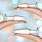 Two hands each holding a piece from the Gläs 3 Piece Anal Training Set by Gläs, showcasing its bottle-shaped design with a flat base and tapered top, perfect for backdoor play, against a soft, colorful bokeh background.